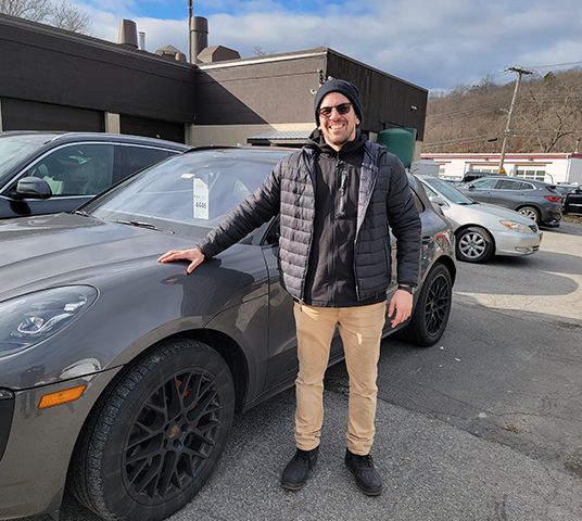 Person standing next to a gray car in a parking lot, dressed warmly.